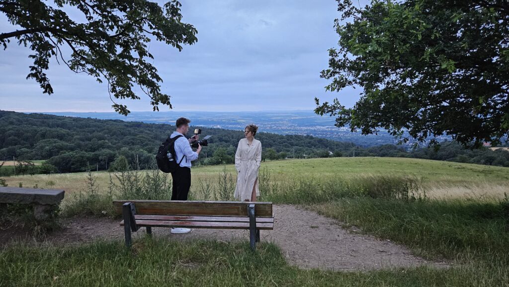 Leandro Matthiessen filmt eine Frau in heller Kleidung auf einer Wiese mit weitem Talblick im Hintergrund
